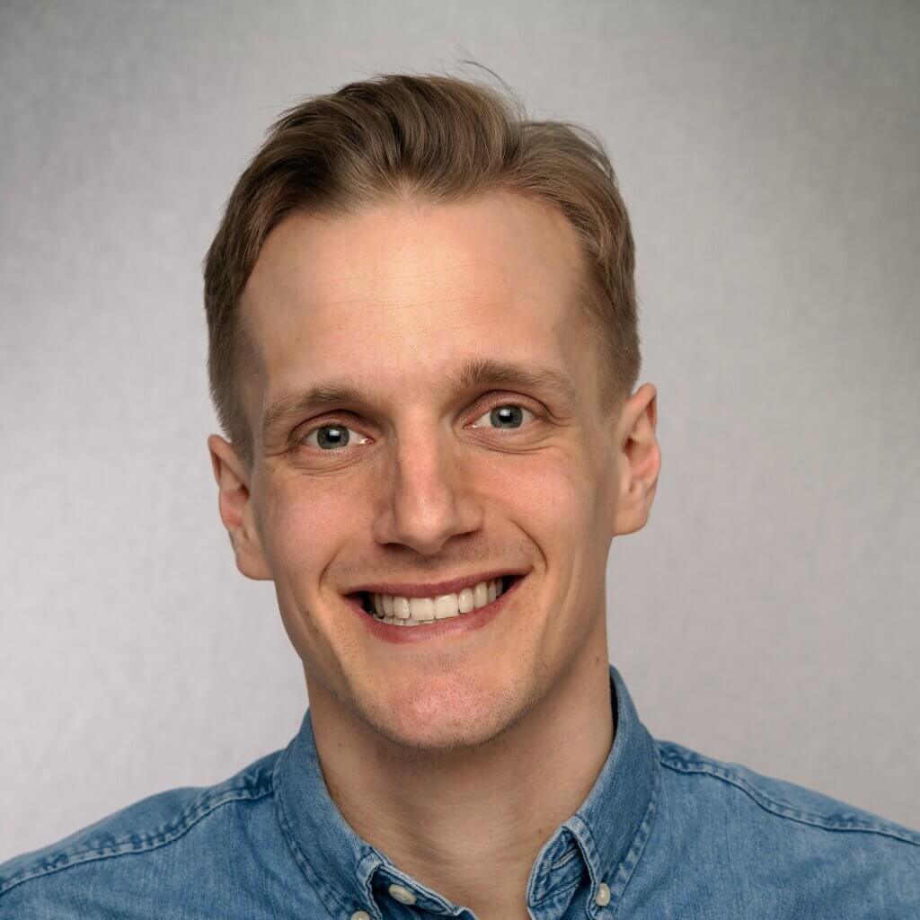 A smiling young man in a denim shirt, looking approachable and friendly. He appears to be a professional in a relaxed setting.