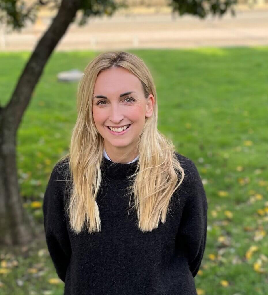 Blonde woman smiling standing in front of a tree on a green lawn