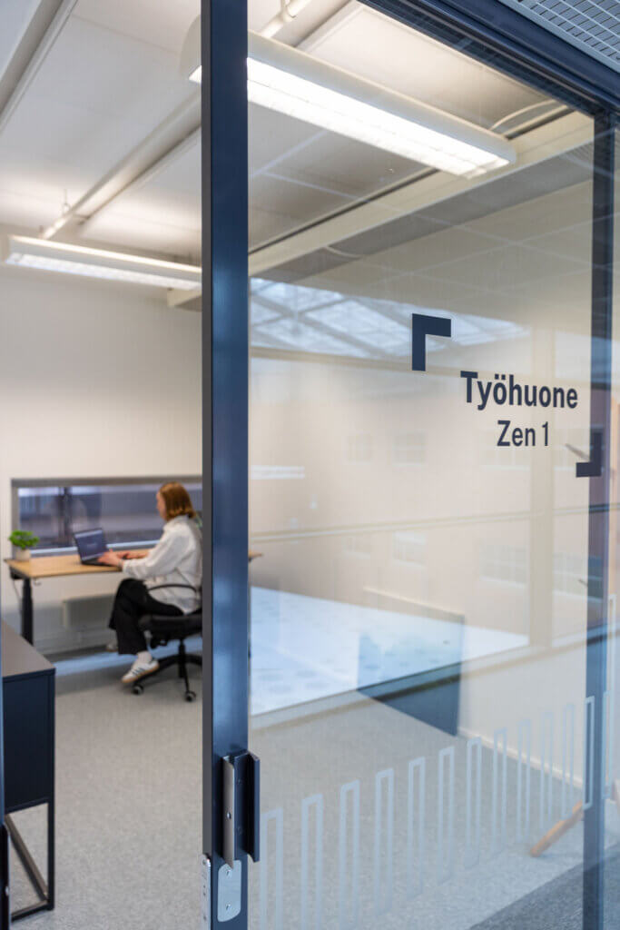 A person sitting at a desk through a glass door with Työhuone Zen 1 written on it