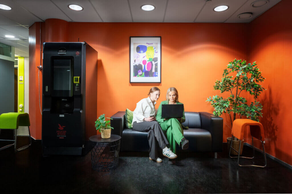 Two business people, one man and one woman, sitting on a couch in an office waiting area with a vending machine nearby.