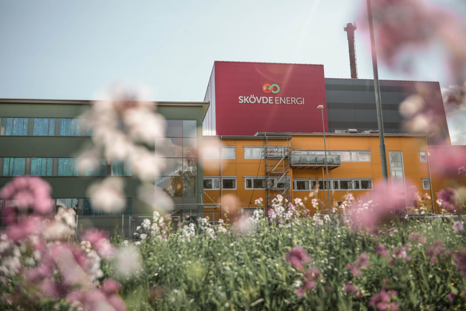 A large industrial building with a red sign, surrounded by flowers, representing a utility or manufacturing facility.