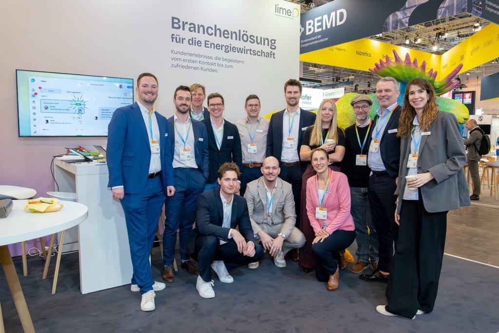 A group of professionals posing for a photo at a business event, with a presentation screen and company information in the background, showcasing a team in a corporate setting.