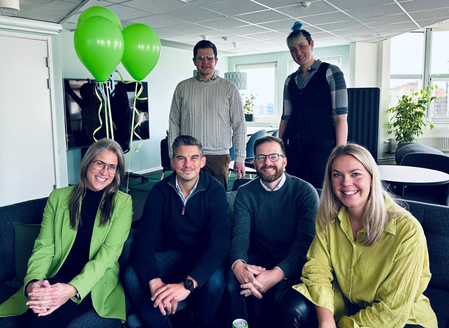 A group of six colleagues smiling and posing together in an office setting with green balloons.