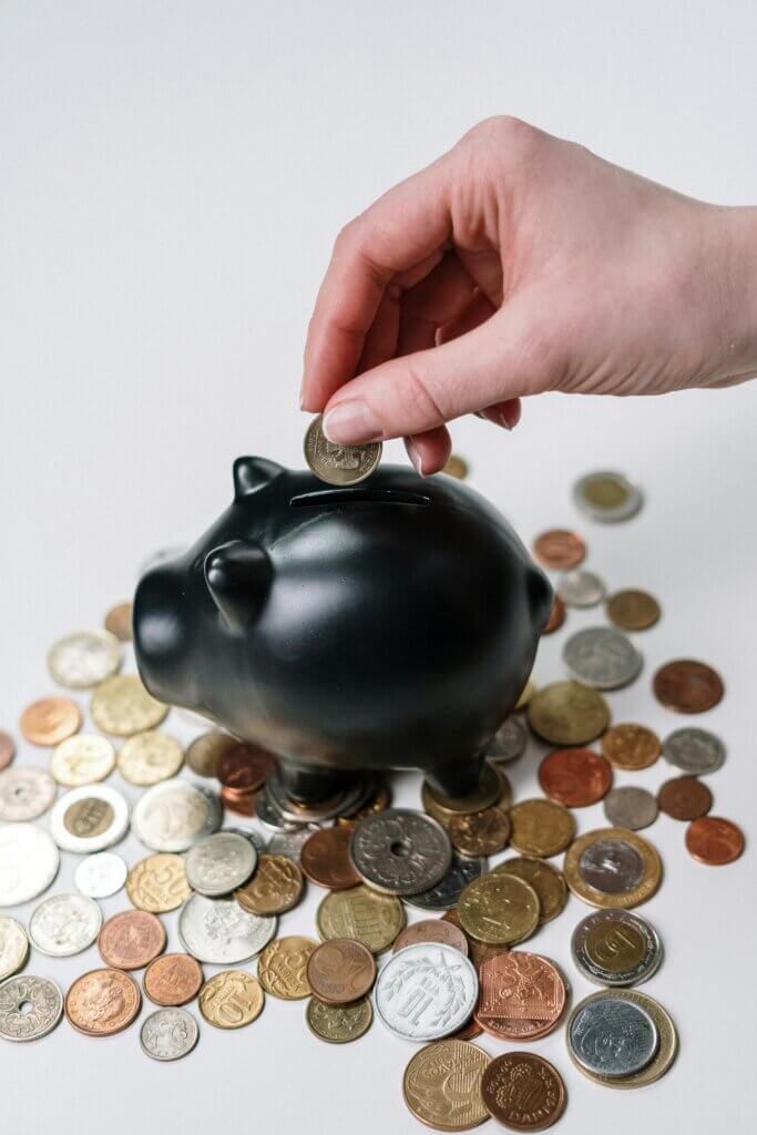 Hand putting a coin into a piggy bank surrounded by various coins, symbolizing savings and financial management, essential for businesses utilizing CRM solutions to optimize their financial operations.
