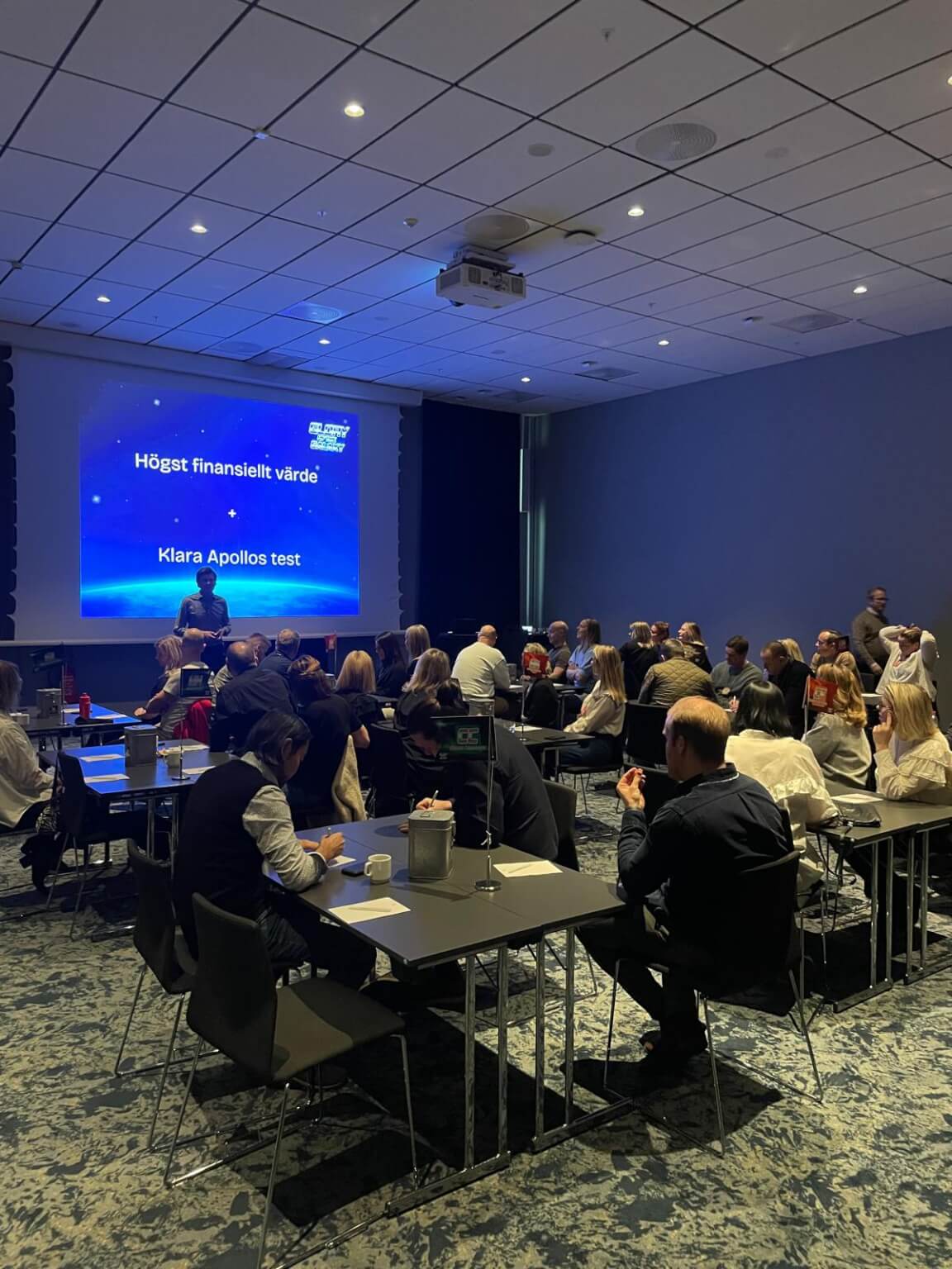A conference room with people seated at tables, attentively listening to a presentation on a large screen.