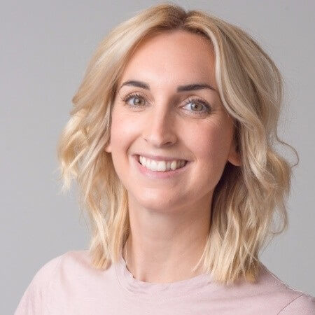 A smiling woman with blond hair looking directly at the camera, wearing a pink top against a plain background, conveying a sense of approachability and professionalism.