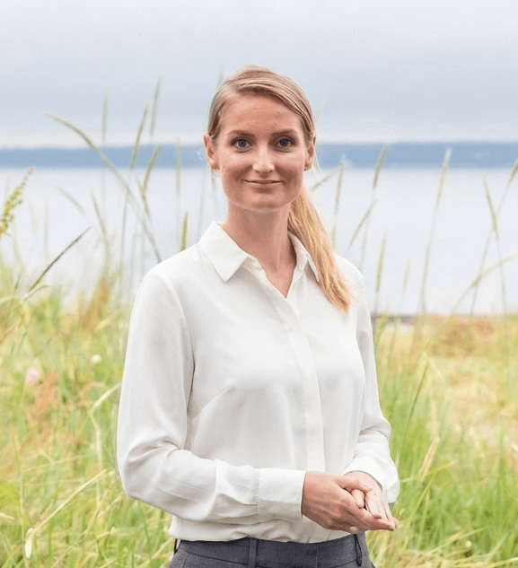 A smiling businesswoman standing in a field of tall grass, representing a professional in a natural setting related to consultancy or real-estate sectors.
