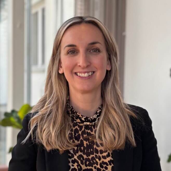 Smiling businesswoman standing in a modern office space, likely a consultant or professional in a CRM industry setting.