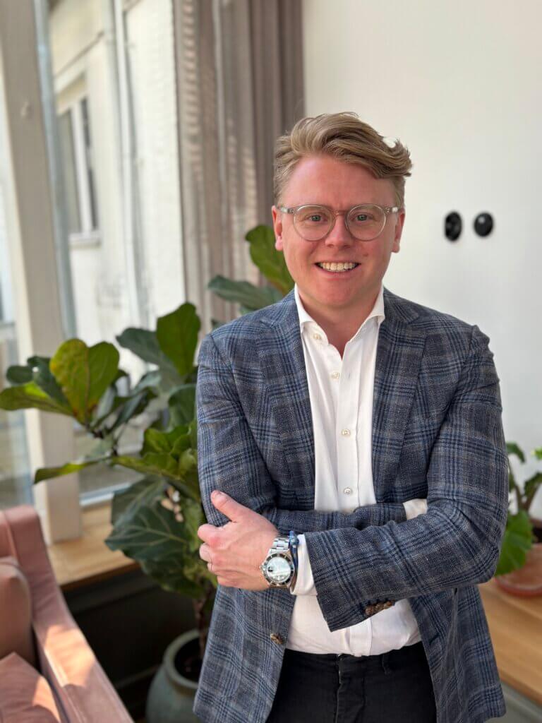 A smiling man in a blue plaid jacket and white shirt standing confidently with his arms crossed in a modern office setting.