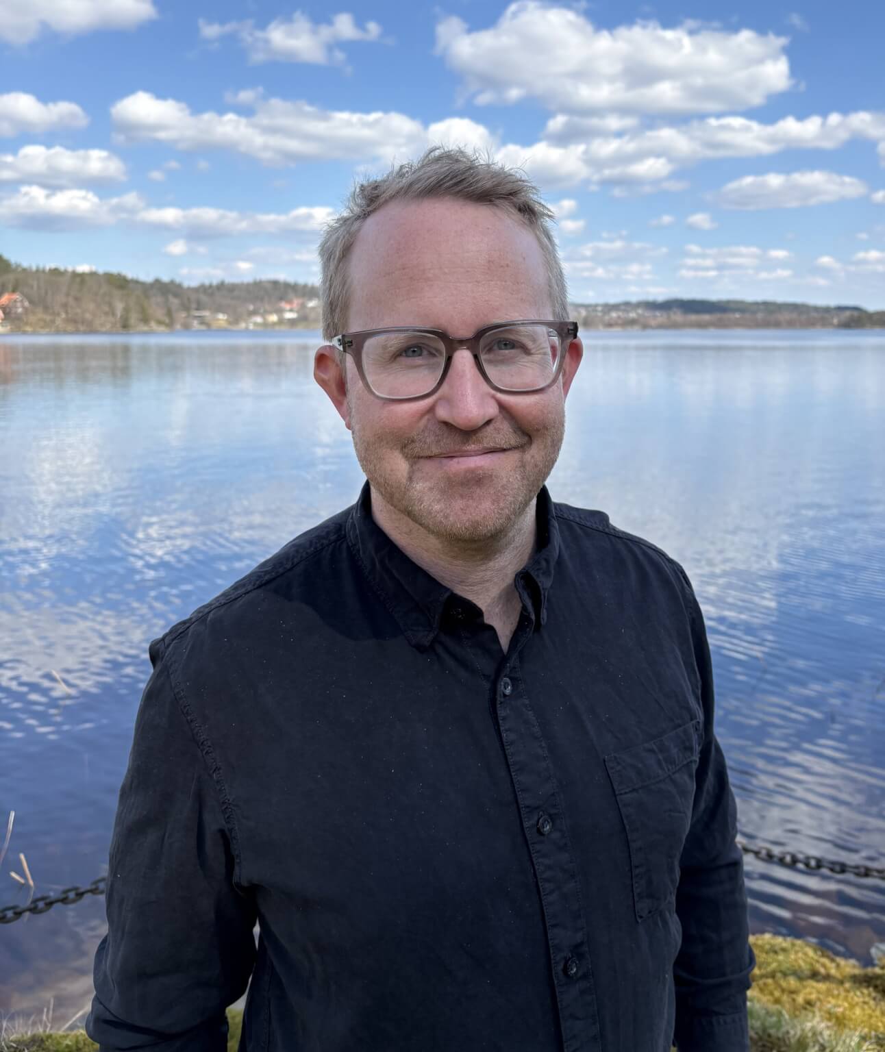 Smiling man in glasses standing by serene lake on a sunny day