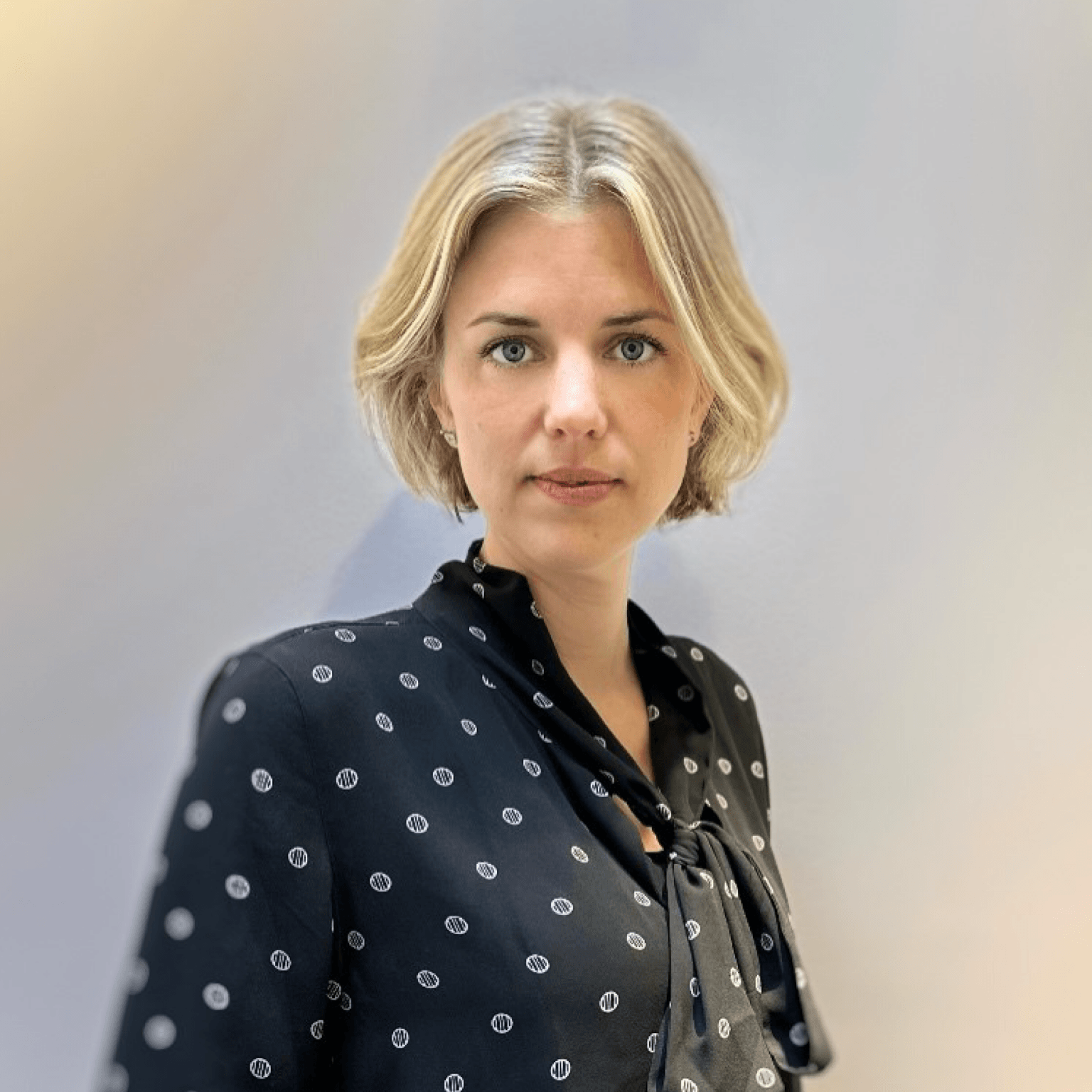 A woman with short blonde hair wearing a black blouse with white polka dots stands confidently, representing professional services in consultancy.