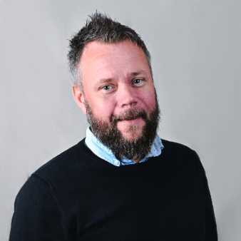 A portrait of a man with a beard looking directly at the camera, wearing a black sweater over a light blue collared shirt.