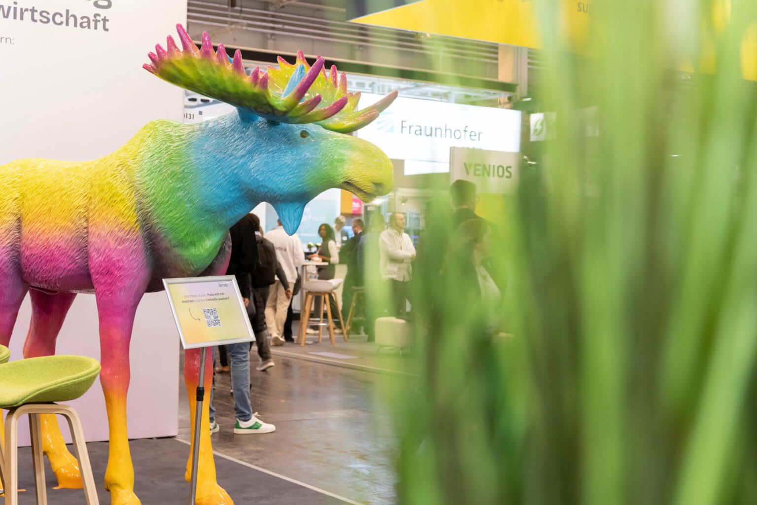 A large, colourful moose sculpture is displayed in an exhibition hall featuring the German word Wirtschaft, evoking a scene related to economic consultancy and business partnerships.