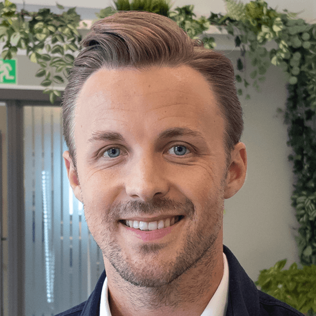 A smiling man with short brown hair and a beard, wearing a blue jacket over a white shirt, in an office setting with green plants in the background.
