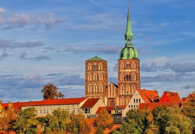 A historic church with red-brick towers and a green spire rises above a tree-lined hillside under a partly cloudy blue sky. The scene captures a scenic European cityscape, suitable for representing municipality or real estate verticals.
