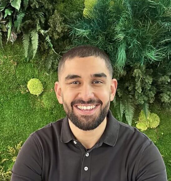 A smiling man in a dark long-sleeved polo shirt seated at a white table, presenting in front of a lush vertical green wall background.